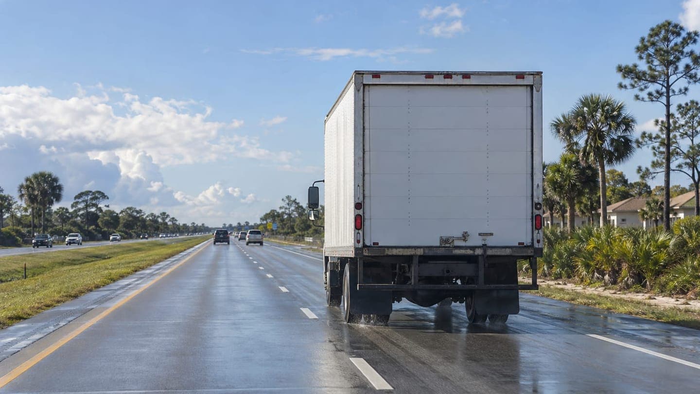 A white moving truck on a wet Florida highway lined with palms and pines.